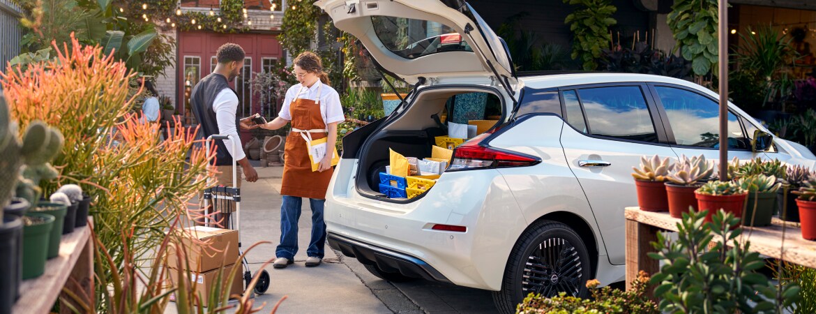 A woman is receiving supplies next to a Nissan Business LEAF vehicle 
