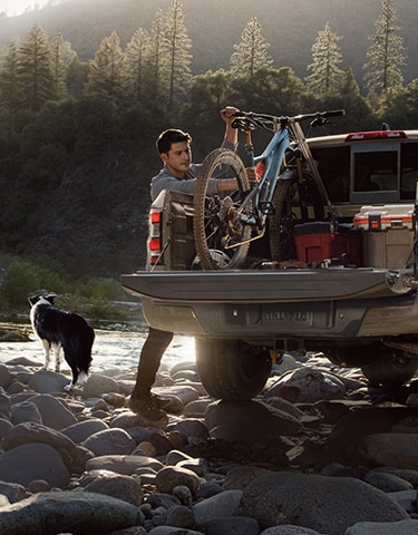 Nissan Frontier 2024 estacionada en la orilla de un arroyo rocoso; el conductor está sujetando una bicicleta y la carga en la parte trasera, mientras su perro mira hacia el arroyo