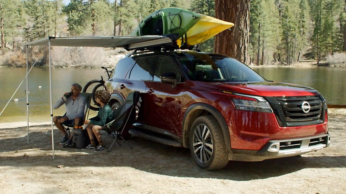 Nissan Pathfinder 2024 al borde del agua, con kayaks encima y una sombrilla retráctil sobre dos personas en sillas de camping.
